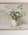 Fine art print framed hanging on a wall. The print shown is a painting of daisies, grasses and leaves in a white vase with a light beige background.