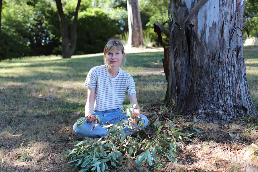 The artist Louise Ernestine Anders is sitting in a grassy field next to a eucalyptus tree holding a branch of eucalyptus leaves