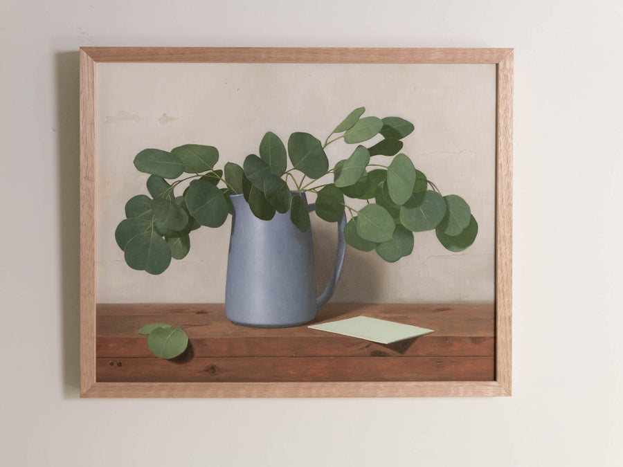 Fine art print framed hanging on a wall.  The print shown is of a painting of eucalyptus leaves in a blue vase with a light background.